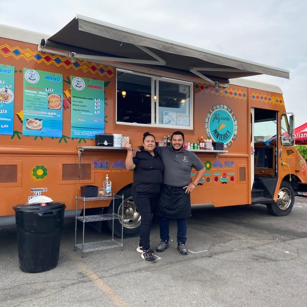 Two smiling Taqueria Monterrey employees stand proudly in front of their orange food truck.