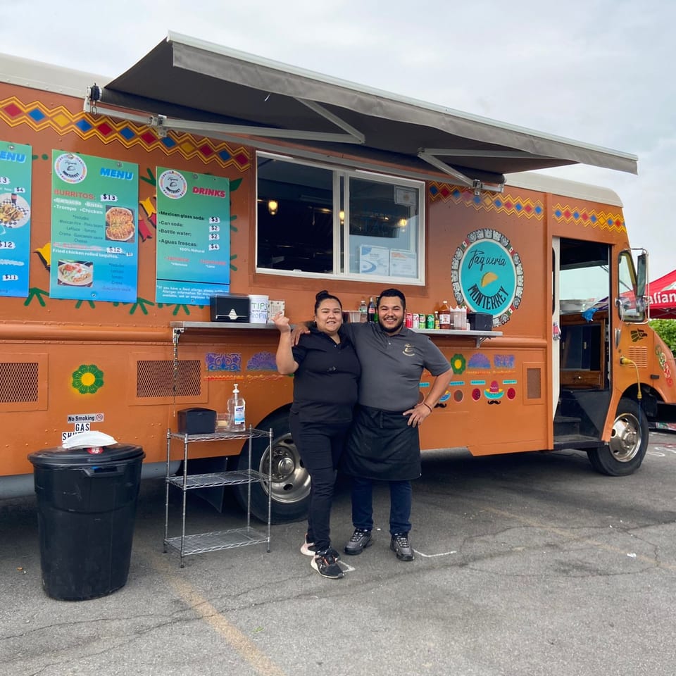 Two smiling Taqueria Monterrey employees stand proudly in front of their orange food truck.