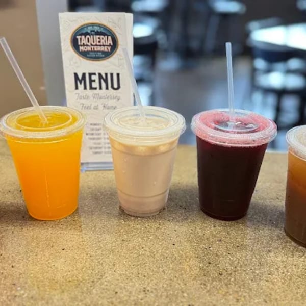 Four colorful drinks in plastic cups with straws on a counter, with a Taqueria Monterrey menu in the background.