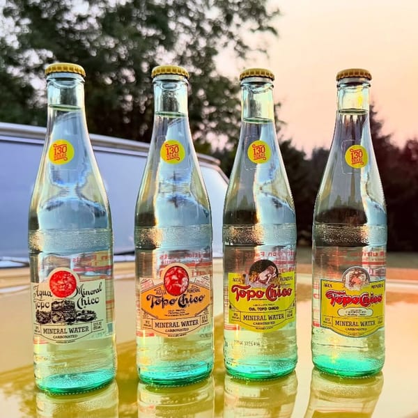 Four bottles of Topo Chico mineral water with yellow caps on a reflective surface outdoors.