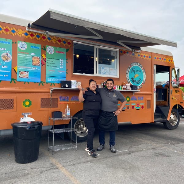 Two smiling Taqueria Monterrey employees stand proudly in front of their orange food truck.