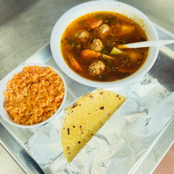 A bowl of meatball soup, a side of rice, and a taco on a metal tray.