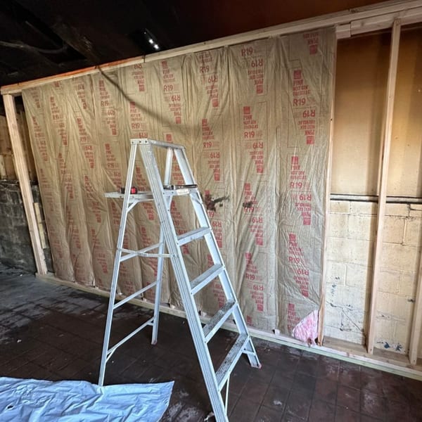 Construction site with insulation on a framed wall, a ladder, and a blue tarp on the floor.