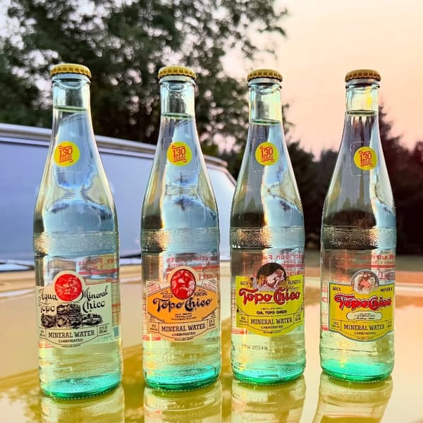 Four bottles of Topo Chico mineral water with yellow caps and labels, on a reflective surface.