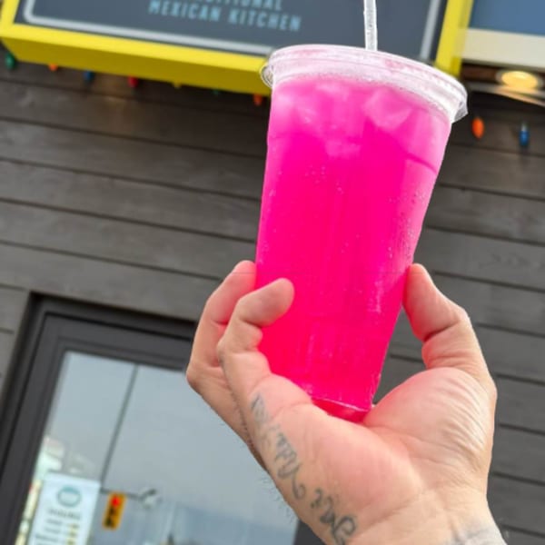 A hand with tattoos holds a bright pink drink with ice and a straw outside Taqueria Monterrey.