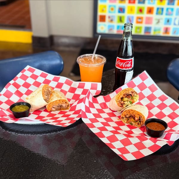 Two burritos on red and white checkered paper, with salsa, a drink, and a Coca-Cola bottle.