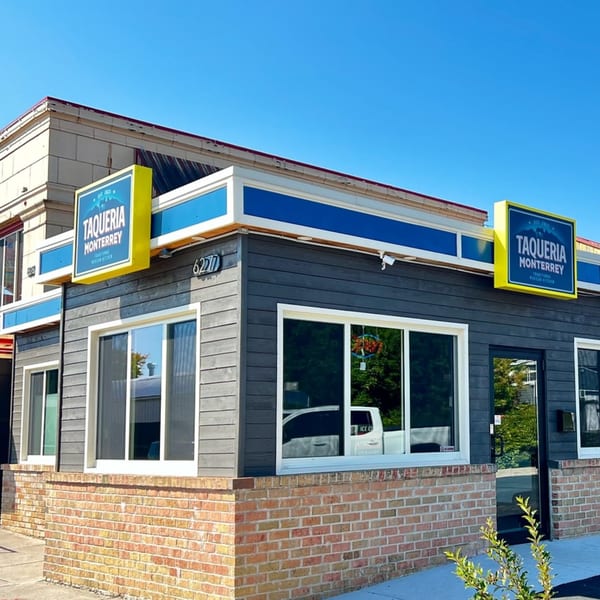 Exterior of Taqueria Monterrey, a gray building with blue and yellow signs, brick base, and large windows.